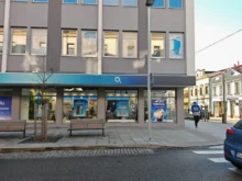 City street storefront with an O2 shop, gray facade, large window posters, and a man in a blue jacket walking nearby.