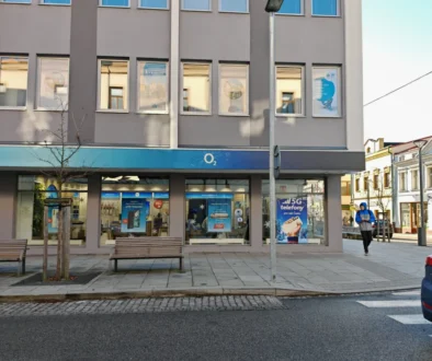 City street storefront with an O2 shop, gray facade, large window posters, and a man in a blue jacket walking nearby.