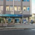 City street storefront with an O2 shop, gray facade, large window posters, and a man in a blue jacket walking nearby.