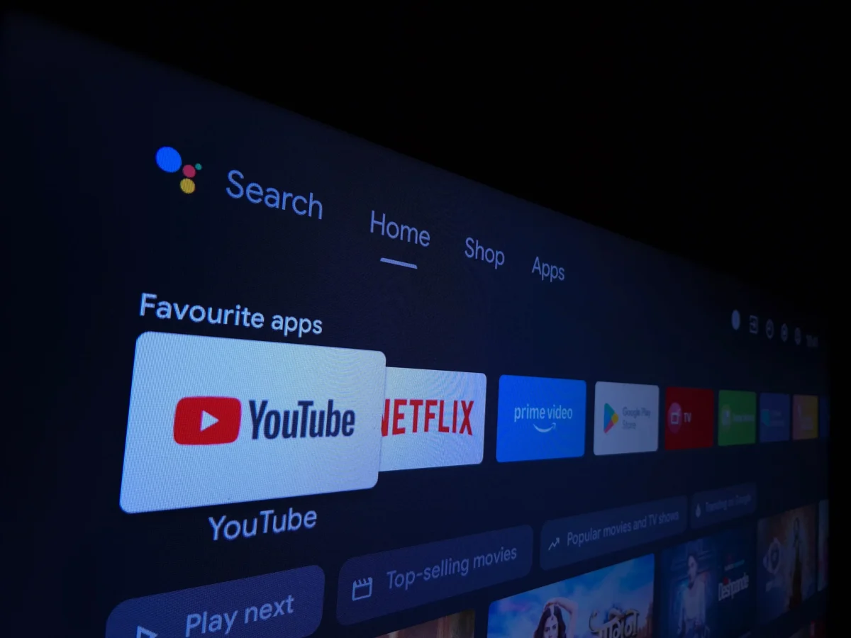 shutterstock_2730156891 1 | TVGURU.cz Smart TV home screen showing a row of apps like YouTube and Netflix with a dark background and blue highlight bar under Search/Home/Shop/Apps navigation.