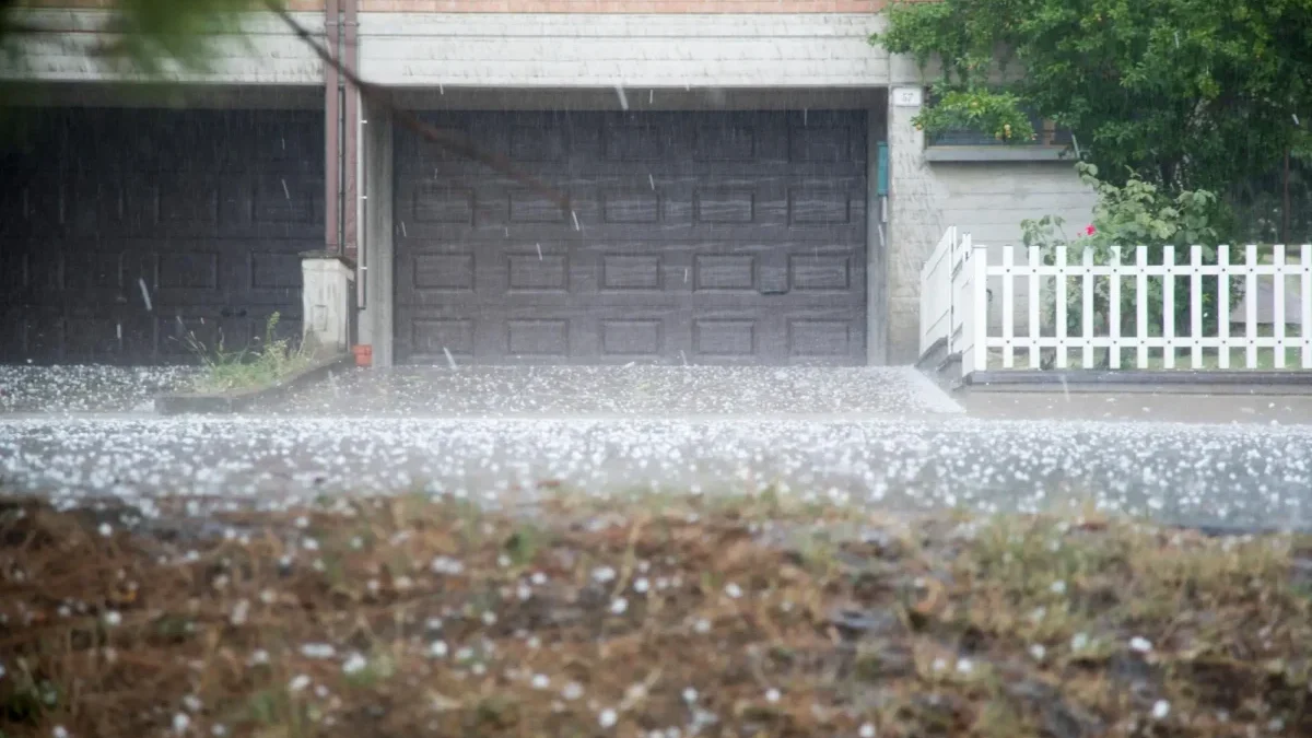 Rainy suburban scene: closed dark garage door beneath a brick facade, white fence on the right, and wet ground with scattered petals.
