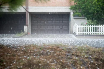 Rainy suburban scene: closed dark garage door beneath a brick facade, white fence on the right, and wet ground with scattered petals.
