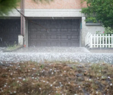 Rainy suburban scene: closed dark garage door beneath a brick facade, white fence on the right, and wet ground with scattered petals.