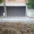 Rainy suburban scene: closed dark garage door beneath a brick facade, white fence on the right, and wet ground with scattered petals.