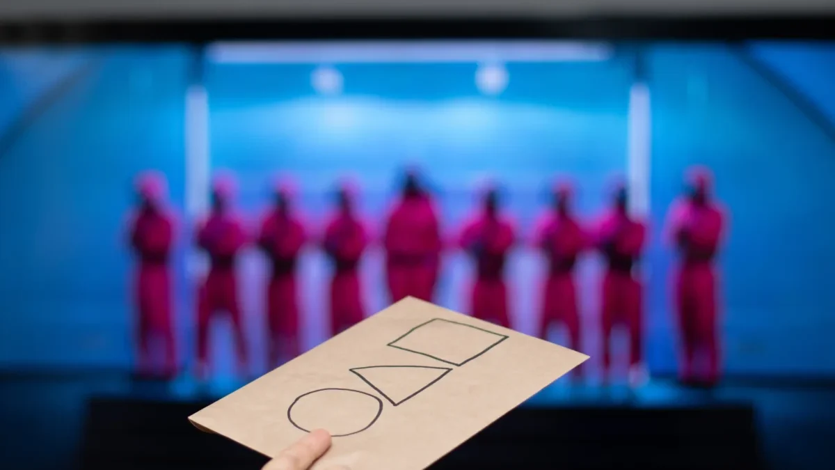 Hand holding a brown card with three empty checkboxes; blurred blue-lit stage with performers in pink outfits in the background.