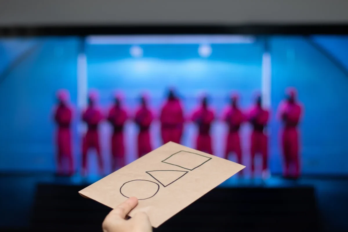 Hand holding a brown card with three empty checkboxes; blurred blue-lit stage with performers in pink outfits in the background.