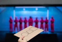 Hand holding a brown card with three empty checkboxes; blurred blue-lit stage with performers in pink outfits in the background.