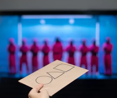 Hand holding a brown card with three empty checkboxes; blurred blue-lit stage with performers in pink outfits in the background.