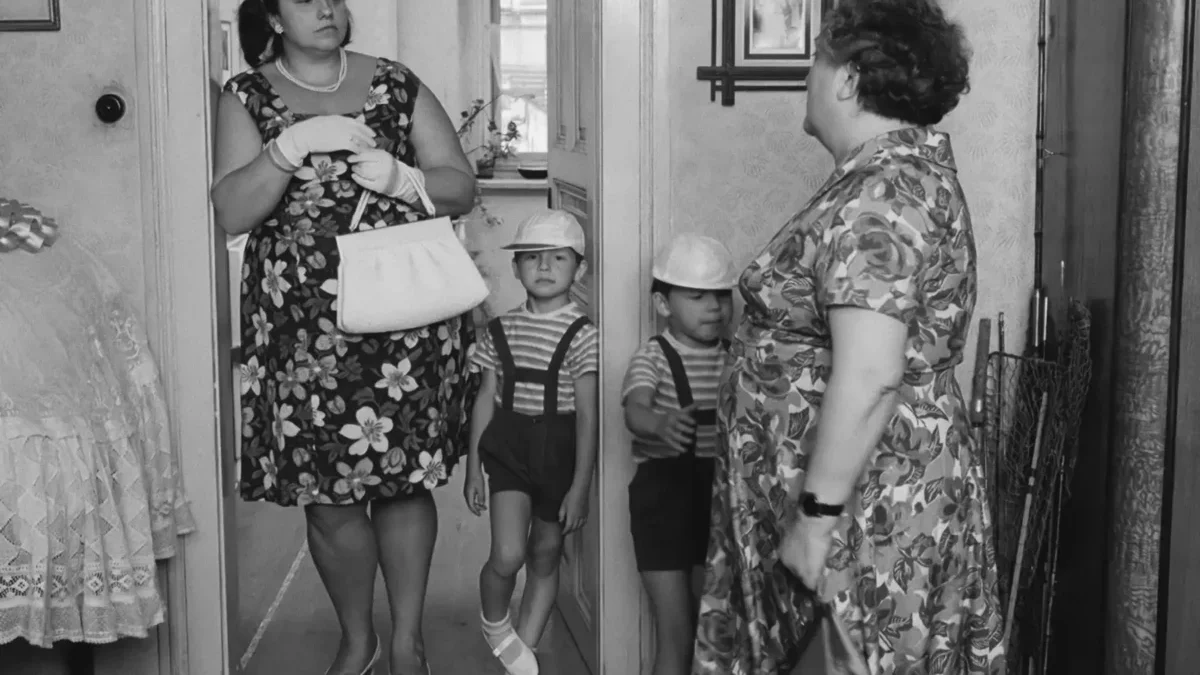 Woman in a floral dress with gloves and pearls stands in a doorway, holding a purse, as two young boys in striped shirts and suspenders stand before her.