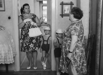 Woman in a floral dress with gloves and pearls stands in a doorway, holding a purse, as two young boys in striped shirts and suspenders stand before her.