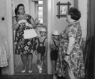 Woman in a floral dress with gloves and pearls stands in a doorway, holding a purse, as two young boys in striped shirts and suspenders stand before her.