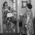 Woman in a floral dress with gloves and pearls stands in a doorway, holding a purse, as two young boys in striped shirts and suspenders stand before her.
