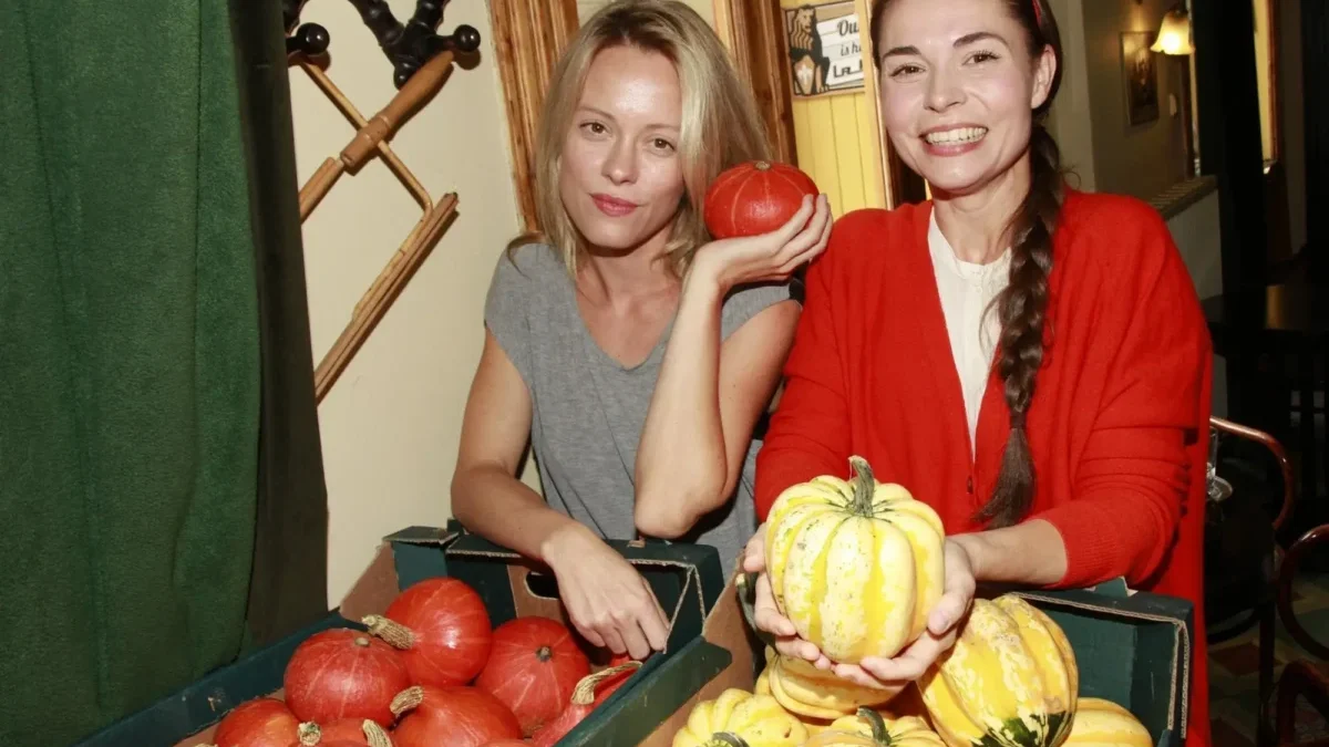 Two women at a pumpkin display, one in gray holding an orange pumpkin while the other in red holds yellow-striped pumpkins and smiles at the camera