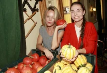 Two women at a pumpkin display, one in gray holding an orange pumpkin while the other in red holds yellow-striped pumpkins and smiles at the camera