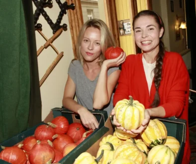 Two women at a pumpkin display, one in gray holding an orange pumpkin while the other in red holds yellow-striped pumpkins and smiles at the camera