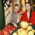 Two women at a pumpkin display, one in gray holding an orange pumpkin while the other in red holds yellow-striped pumpkins and smiles at the camera