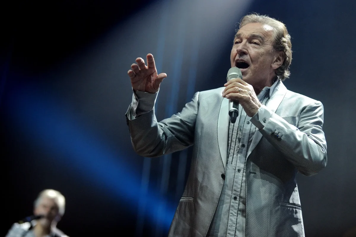 Se seriálem je spojen i zpěvák Karel Gott. Zdroj: Shutterstock | TVGURU.cz Male singer in a gray suit sings into a handheld microphone on a dark stage with blue lighting