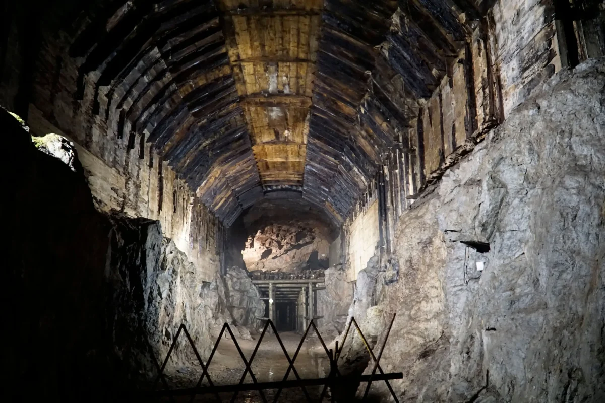 nacisti | TVGURU.cz Podzemní tunel s klenutými dřevěnými podpěrami a drsnými skalními stěnami, bariéra blokující cestu v popředí.