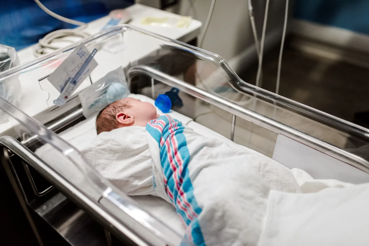 Newborn baby wrapped in a blue and pink striped blanket lying in a clear hospital bassinet with metal rails.