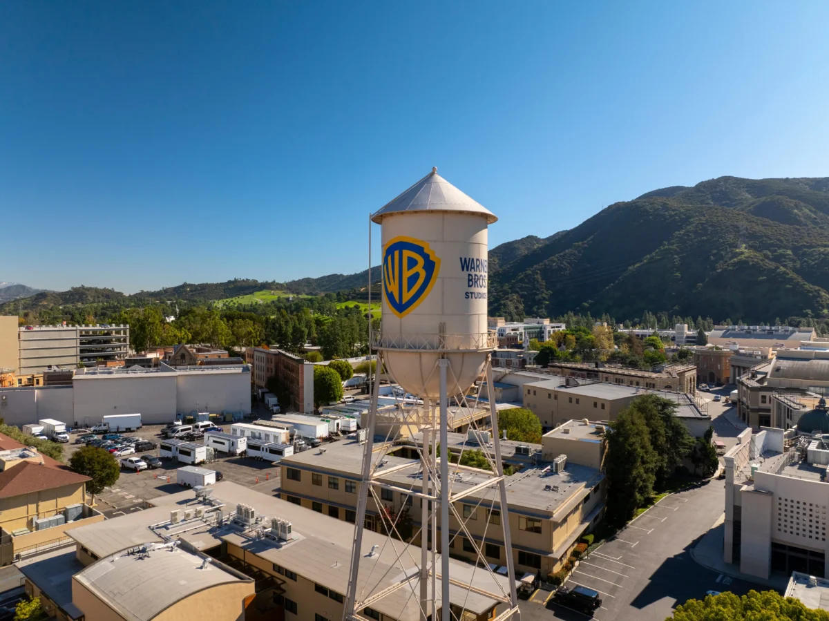 shutterstock_2713330203 1 | TVGURU.cz Water tower with the Warner Bros. logo rising above a suburban industrial area with hills in the distance under a clear blue sky.60 characters