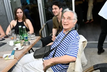 dva muži a žena sedící u jednoho stolu. nejbližší muž je postarší a má na sobě modro bílé pruhované triko a černou brašnu přes rameno, vedle něho sedí mladší muž s černými vlasy a zeleným trikem a po jeho pravici sedí mladá burneta s černým tílkem