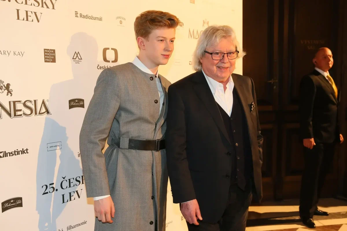 Two men posing on a red carpet at a film event, one in a gray suit with a belt and the other in a black suit, backdrop with sponsor logos.