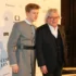 Two men posing on a red carpet at a film event, one in a gray suit with a belt and the other in a black suit, backdrop with sponsor logos.