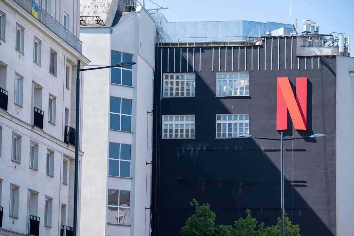 shutterstock_2318307591 1 | TVGURU.cz White multi-story residential buildings on the left beside a dark wall with a large red letter N on the right, under a clear blue sky.