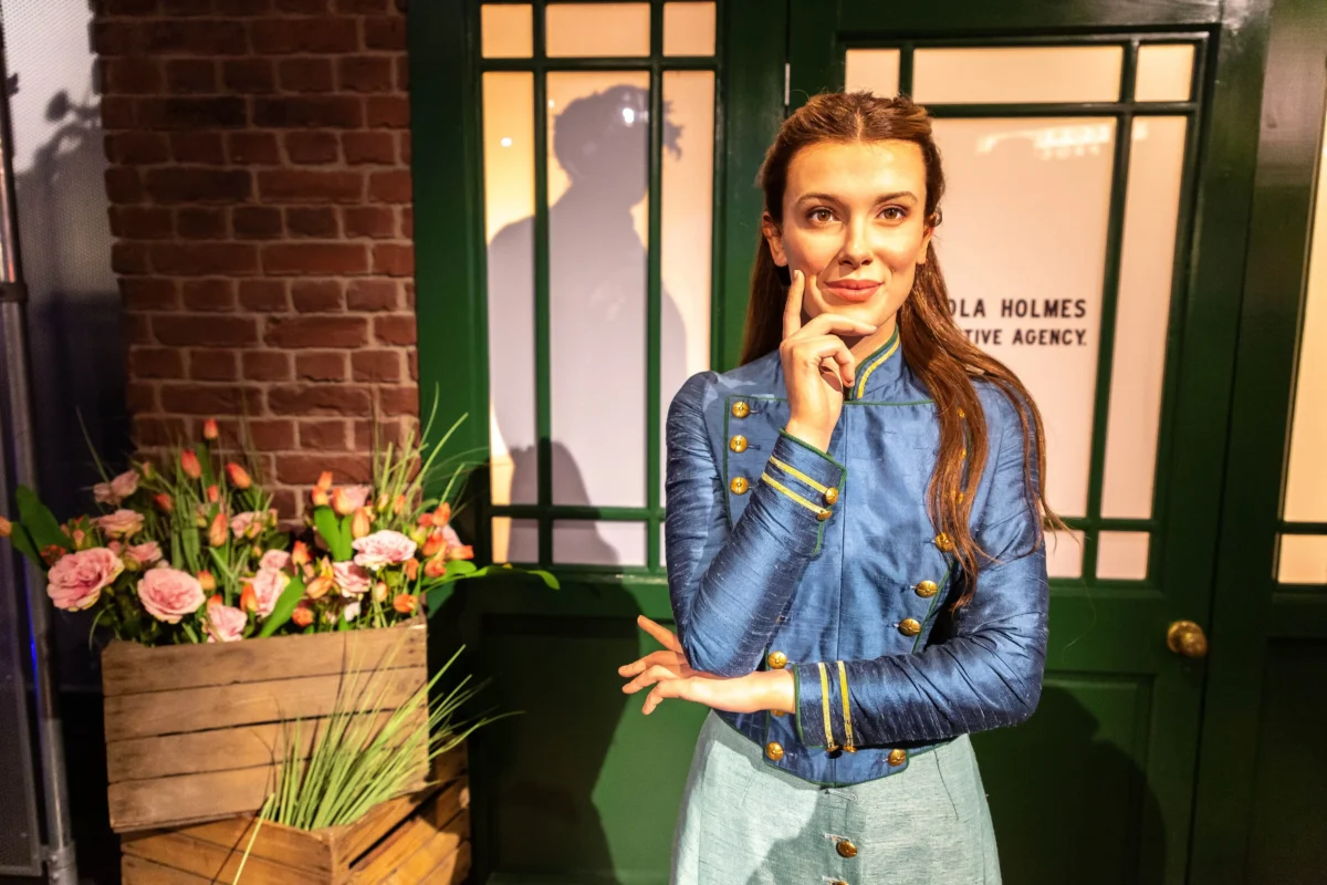 shutterstock_2668034627 1 | TVGURU.cz Woman in a blue military-style jacket poses with a thoughtful expression, hand to chin, in front of a green-framed window and brick wall with flowers nearby.