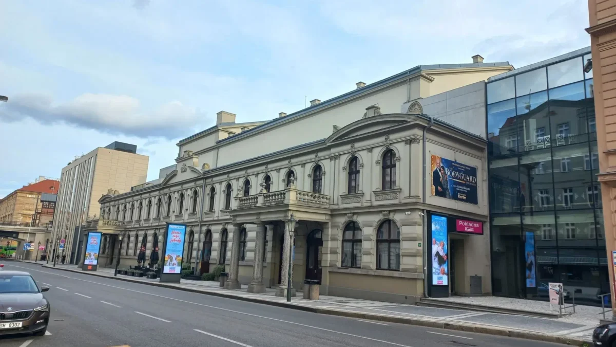 Historic beige building with arched windows and decorative cornices along a city street; modern glass building to the right; blue sky above.