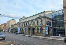 Historic beige building with arched windows and decorative cornices along a city street; modern glass building to the right; blue sky above.