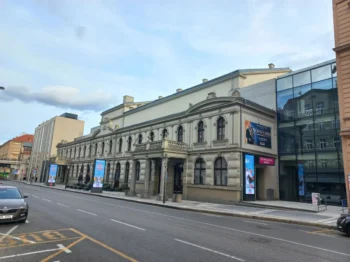 Historic beige building with arched windows and decorative cornices along a city street; modern glass building to the right; blue sky above.