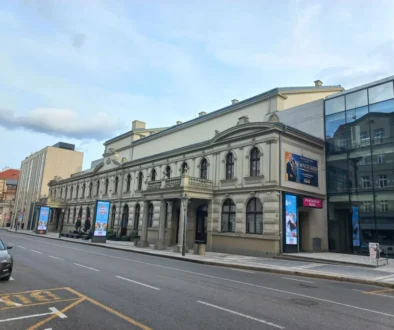 Historic beige building with arched windows and decorative cornices along a city street; modern glass building to the right; blue sky above.