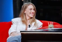Smiling woman in a white blazer with a lapel mic, seated on a red sofa during an interview setting.