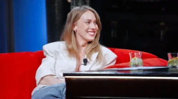Smiling woman in a white blazer with a lapel mic, seated on a red sofa during an interview setting.
