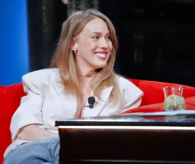 Smiling woman in a white blazer with a lapel mic, seated on a red sofa during an interview setting.