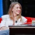 Smiling woman in a white blazer with a lapel mic, seated on a red sofa during an interview setting.