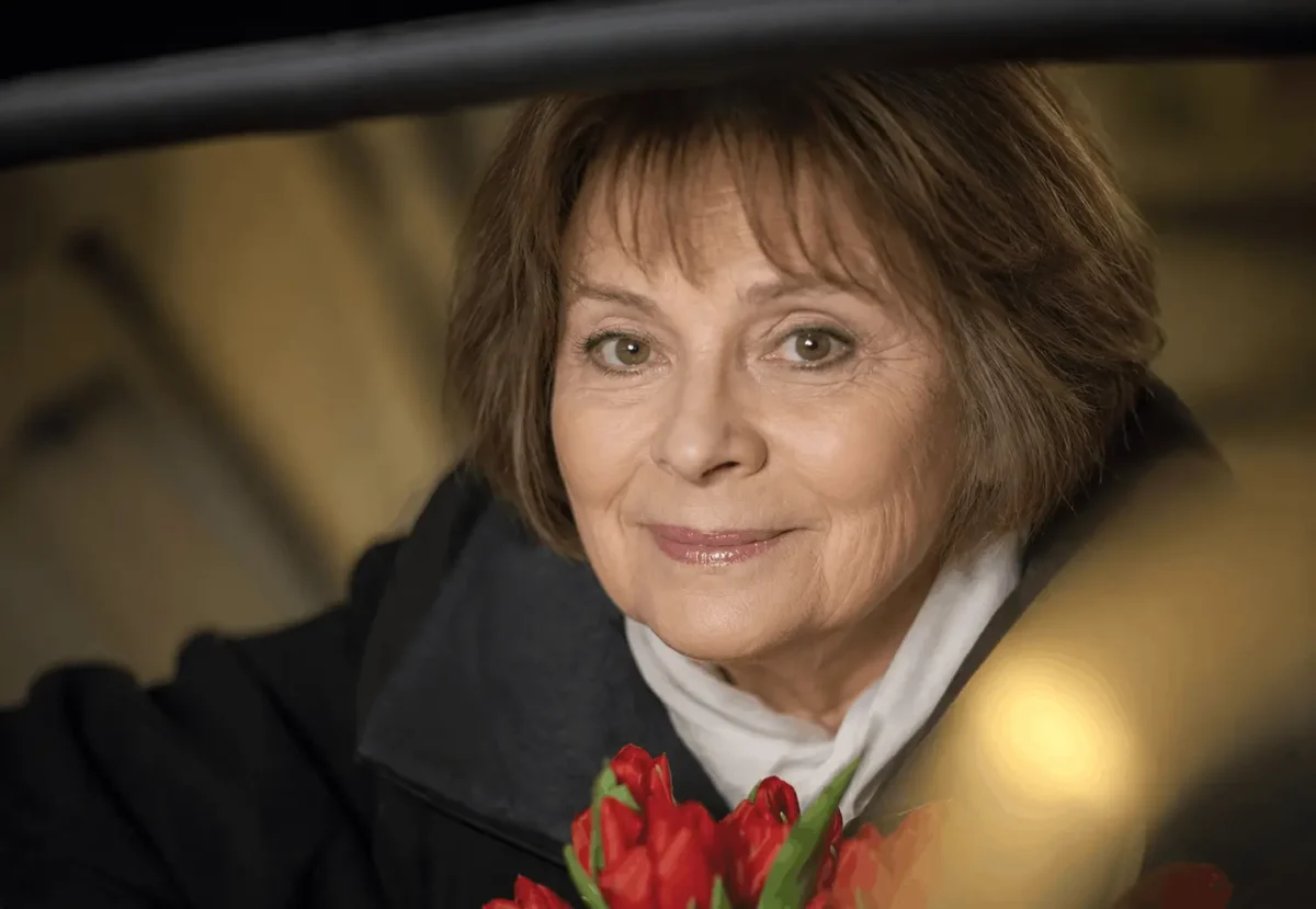 ChatGPT Image 17 6 2026 16_15_29 | TVGURU.cz Smiling elderly woman in a dark coat holds a bouquet of red tulips, portrait-style close-up inside a dim setting.