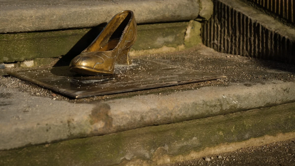 shutterstock_1618525009 1 | TVGURU.cz Old bronze shoe sculpture resting on a wooden board on a mossy stone staircase outdoors, with dirt around it.
