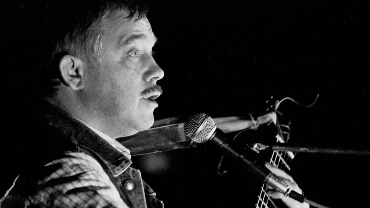 Profile of a male musician with a mustache singing into a microphone while playing an acoustic guitar on stage (black and white).| Informative image with musician performing.