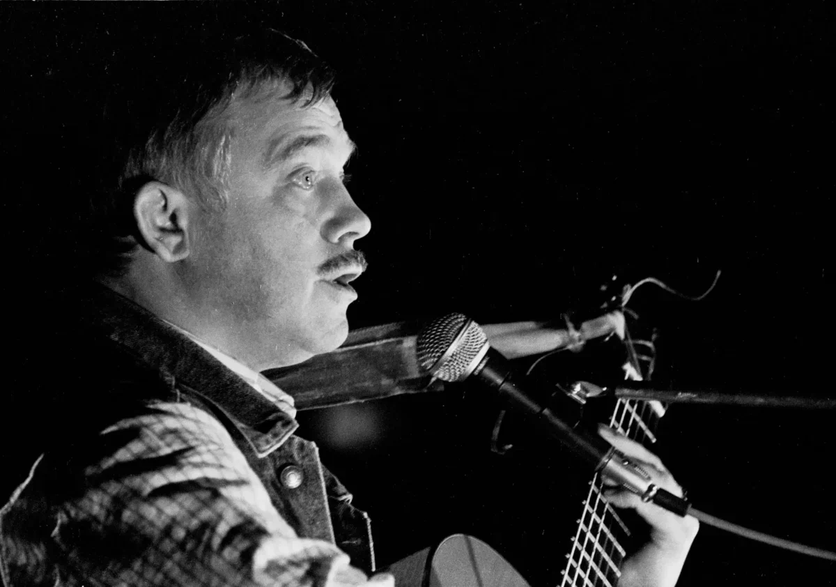 Profile of a male musician with a mustache singing into a microphone while playing an acoustic guitar on stage (black and white).| Informative image with musician performing.