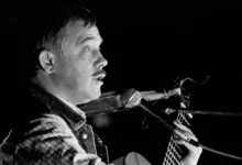 Profile of a male musician with a mustache singing into a microphone while playing an acoustic guitar on stage (black and white).| Informative image with musician performing.