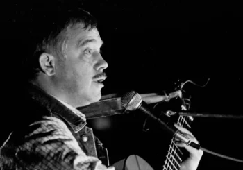 Profile of a male musician with a mustache singing into a microphone while playing an acoustic guitar on stage (black and white).| Informative image with musician performing.