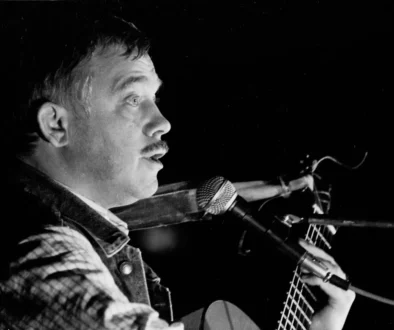 Profile of a male musician with a mustache singing into a microphone while playing an acoustic guitar on stage (black and white).| Informative image with musician performing.
