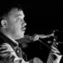 Profile of a male musician with a mustache singing into a microphone while playing an acoustic guitar on stage (black and white).| Informative image with musician performing.