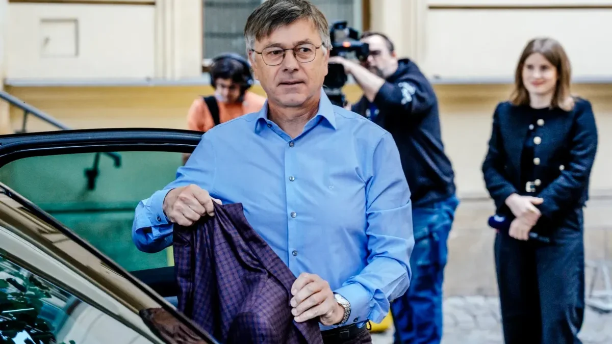 Man in a light blue shirt exits a car, holding a dark plaid blazer as photographers and a woman watch in the background.