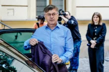 Man in a light blue shirt exits a car, holding a dark plaid blazer as photographers and a woman watch in the background.