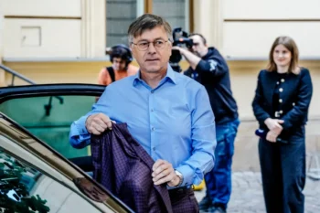 Man in a light blue shirt exits a car, holding a dark plaid blazer as photographers and a woman watch in the background.