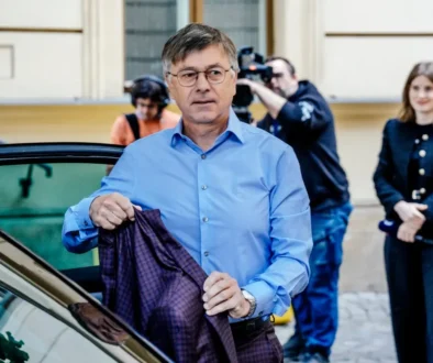 Man in a light blue shirt exits a car, holding a dark plaid blazer as photographers and a woman watch in the background.