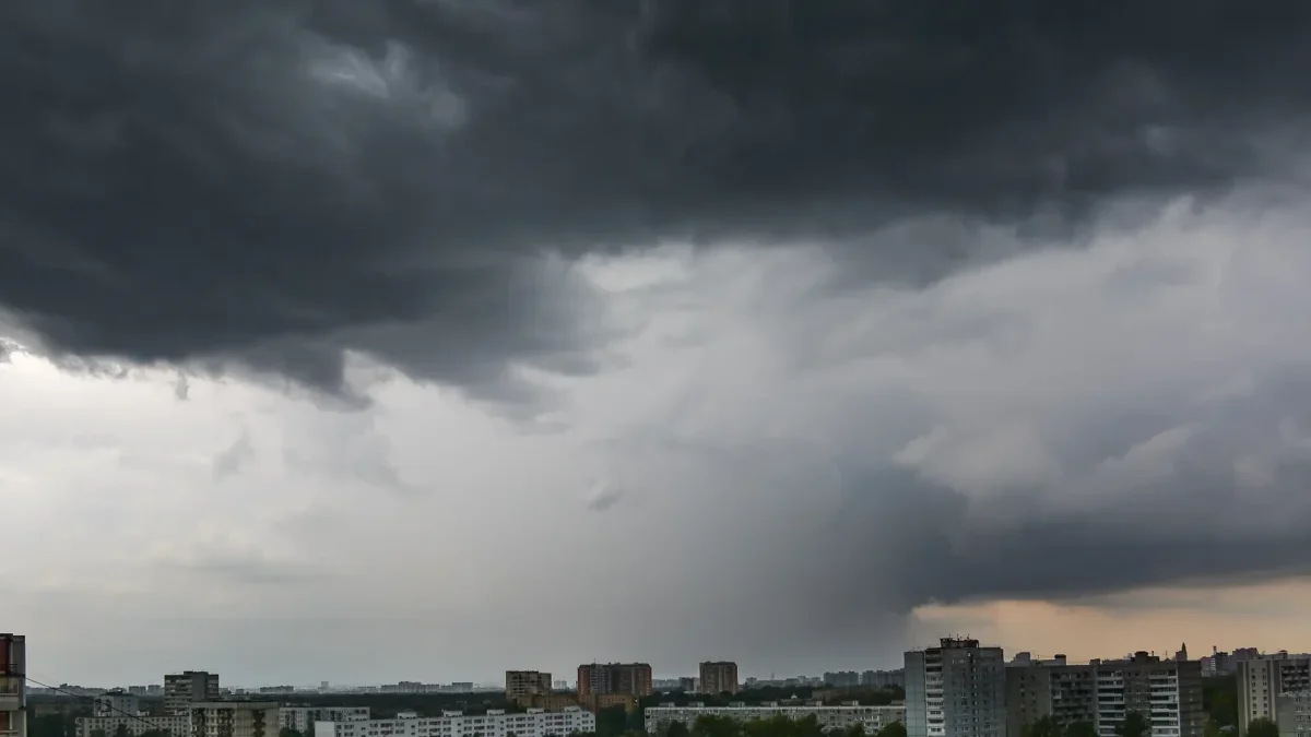 Dark storm clouds loom over a city skyline, with apartment blocks and trees below at dusk/light clouded horizon.